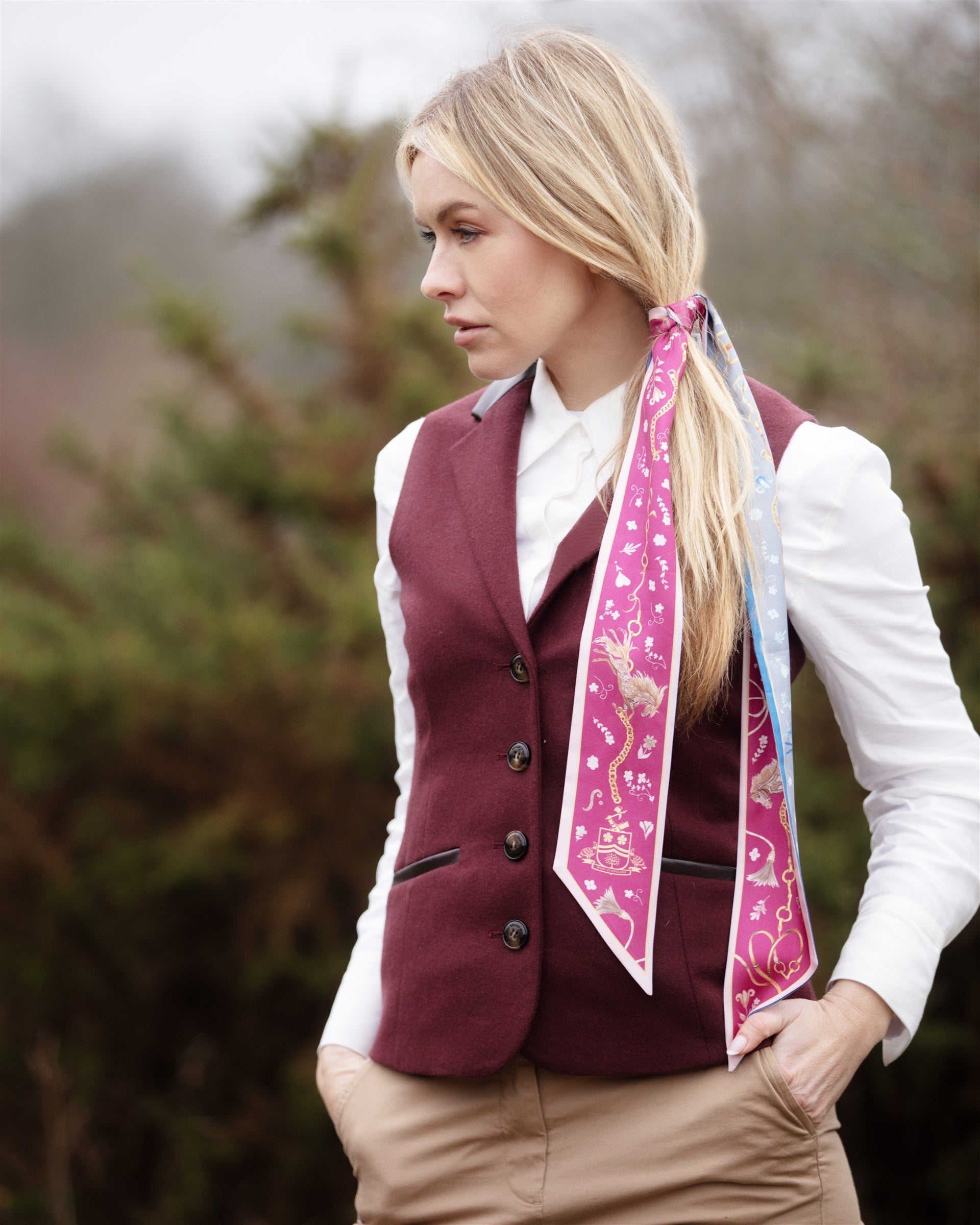 Woman wearing a small silk twilly scarf in pink and pale blue with delicate country-inspired detailing, styled as a hair accessory with a burgundy waistcoat and white shirt outdoors.