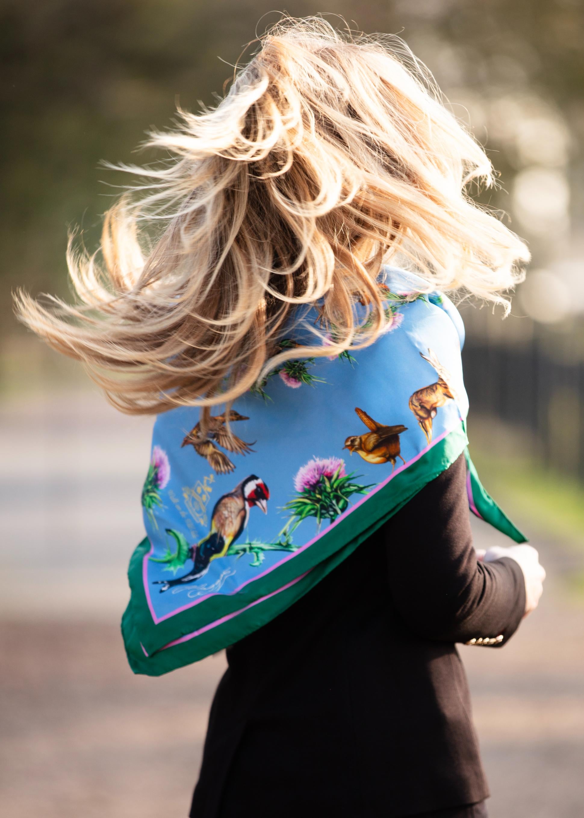 Woman wearing a ladies large square silk scarf in cobalt blue with detailed British wildlife and country-inspired artwork, styled across the back as a statement silk scarf outdoors.