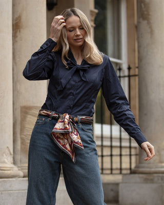 Woman wearing a navy blue blouse with jeans and black scarf belt standing in front of a building.