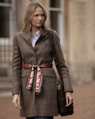 Woman wearing a brown coat with a leather scarf belt and carrying a bag, standing outdoors.