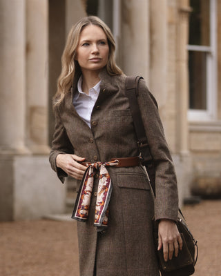 Woman in a brown coat with a patterned leather scarf belt standing in front of a building.