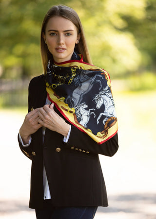 A woman wearing a black blazer and a black silk scarf with a detailed graphic print of horses and gold chains, standing outdoors against a blurred green background.