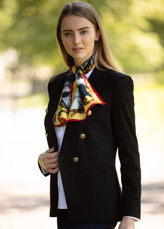 A woman wearing a black double-breasted blazer with gold buttons, a white shirt, and a patterned silk scarf with red, yellow, and black details, standing outdoors against a blurred green background.