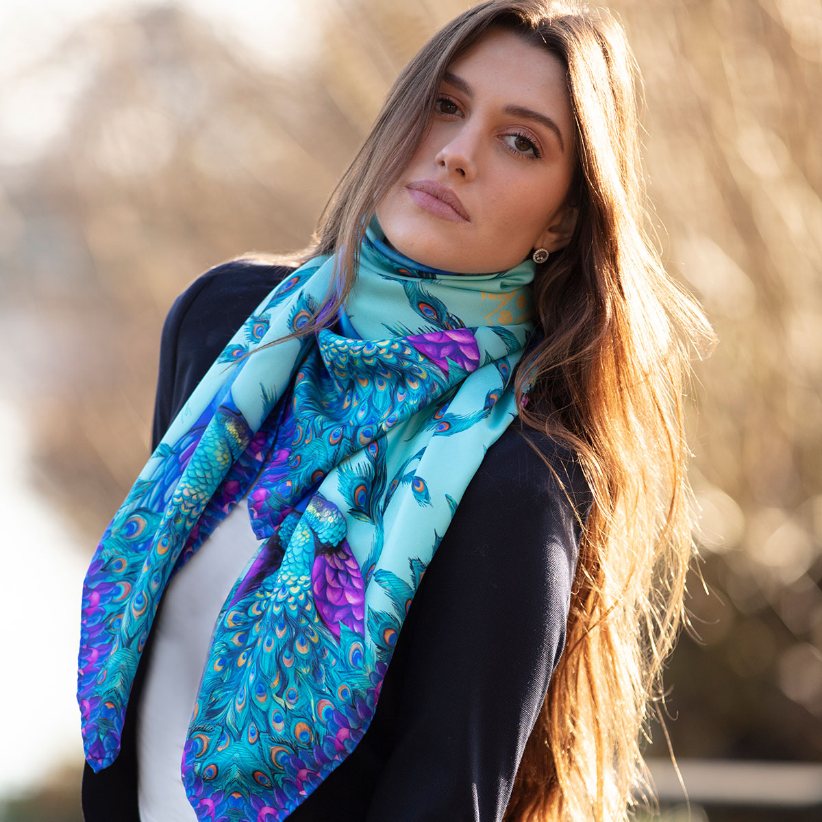 Ladies large square silk scarf in bright turquoise with colourful peacock and feather design in blue and purple, worn as a statement neck scarf with a tailored navy blazer.
