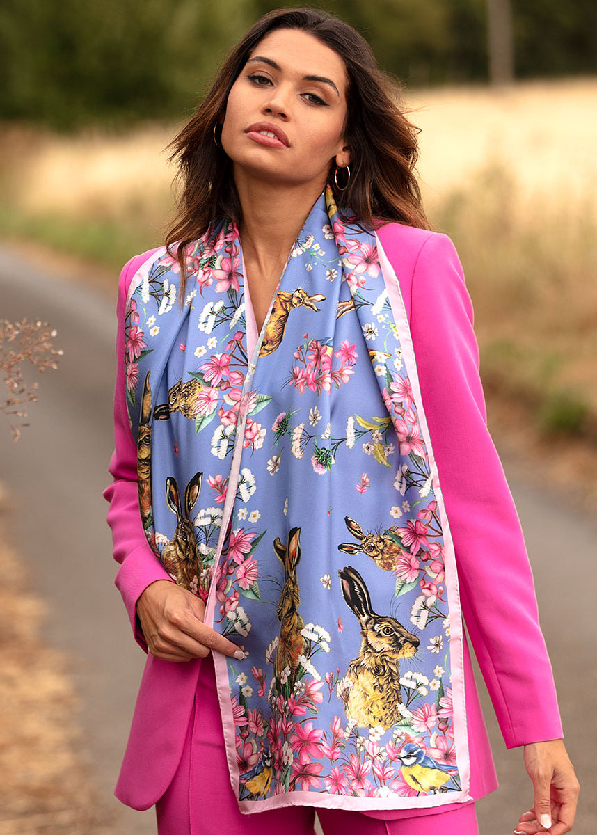 Ladies classic silk scarf in periwinkle blue with pink floral, hare and wildlife design, worn elegantly over a bright pink tailored suit outdoors.