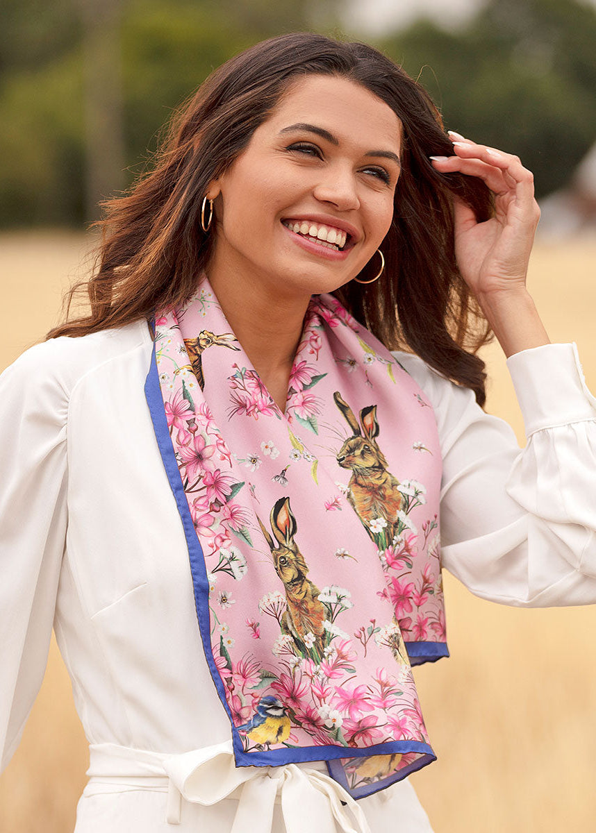 Ladies narrow silk scarf in blush pink with hare, wildflower and British wildlife design, worn as a long neck scarf with a white dress outdoors.